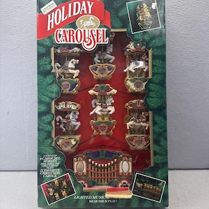 May include: A Holiday Carousel musical decoration box featuring six individual carousel horses and a circus organ. The packaging displays green pine branch borders, red accents, and text stating A string of 6 carousel horses ride while the circus organ plays 21 favorite Christmas carols. Lighted musical carousel hear them play.