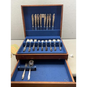 May include: A wooden flatware chest with a blue velvet interior, containing a set of silver-toned knives, forks, and spoons. The top section holds upright knives and horizontal forks and spoons, while the bottom drawer contains two additional utensils.