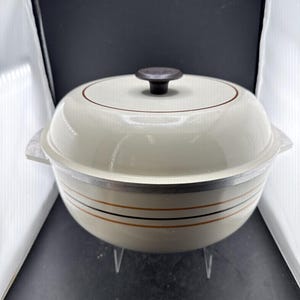 May include: A round, off-white Dutch oven with a matching lid and a dark brown knob. The pot features a metallic rim and horizontal stripes in brown and black around the centre. It has integrated side handles and a smooth, glossy finish.