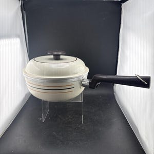 May include: A cream-colored saucepan with a matching lid, featuring horizontal brown and orange stripes around the exterior. The pan has a long, black handle with a small metal loop at the end and a black knob on the center of the lid.