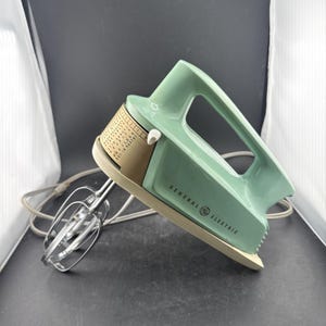May include: A vintage General Electric hand mixer in a light mint green and beige color scheme. The appliance features a top handle, a speed control dial with printed text, and two metal beaters attached to the front. A white power cord is included.