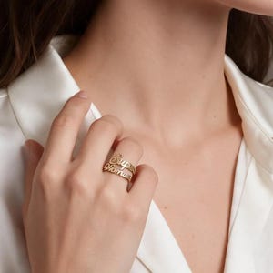 Può includere: Un anello color oro caratterizzato da due fasce sovrapposte con scritte in corsivo. L'anello è indossato su un dito, mettendo in risalto la finitura metallica e il design intricato e fluido del testo integrato nella struttura.