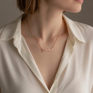 May include: A rose gold-toned metal necklace featuring a delicate chain and a central nameplate pendant with the name Emma written in a cursive script font. The necklace rests against the collarbone, showcasing a polished finish and simple design.