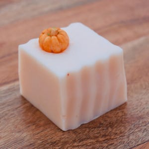 May include: A square-shaped off-white bar of soap with a smooth, flat top and vertical fluted sides. A small, detailed orange pumpkin figurine is embedded into the top surface of the soap, positioned near the left corner of the square block.