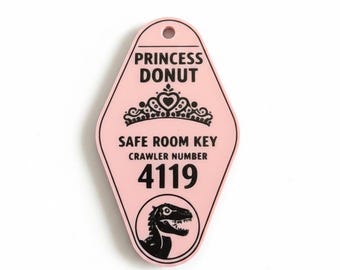May include: A light pink, diamond-shaped motel-style keychain featuring black text that reads PRINCESS DONUT, SAFE ROOM KEY, CRAWLER NUMBER 4119. The design includes a black graphic of a tiara above the text and a dinosaur silhouette below.