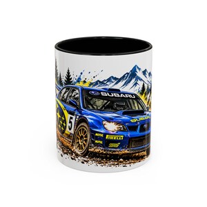 May include: A white ceramic mug with a black interior featuring a graphic of a blue Subaru rally car with the number 5, yellow accents, and the text Subaru, Motul, Pirelli, and STI, set against a backdrop of mountains and pine trees.