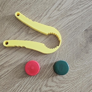 May include: A yellow plastic jar opener tool with a serrated inner grip and two handles, accompanied by one solid red circular bottle cap and one dark green circular bottle cap featuring the printed text L4066 082027 on its top surface.