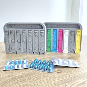 May include: Two vertical pill organizers labeled One Week Pill Organizer with seven columns each. One is solid gray, the other features multicolored columns in green, blue, pink, white, gray, and yellow. Three blister packs of capsules lie in front.