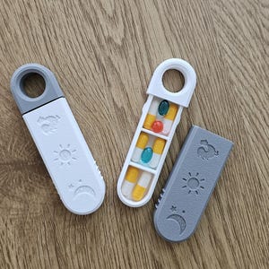 Peut inclure: Deux piluliers avec une poignée en anneau, l'un blanc et l'autre gris, présentant des icônes en relief d'un coq, d'un soleil et d'une lune. Un pilulier est fermé, tandis que l'autre est ouvert, révélant quatre compartiments avec des gélules.