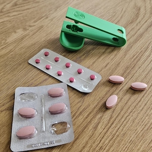 Pode incluir: Um cortador de comprimidos de plástico verde posicionado perto de duas embalagens blister prateadas contendo pequenos comprimidos ovais cor-de-rosa, com três comprimidos ovais cor-de-rosa adicionais soltos na superfície, mostrando o dispositivo.