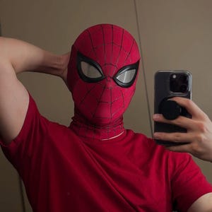 Könnte beinhalten: Eine rote Spider-Man-Maske mit schwarzem Spinnennetz-Muster und großen, weißen, mandelförmigen Augenlinsen mit dicken schwarzen Rändern. Die Maske besteht aus strukturiertem Stoff und wird mit einem schlichten roten T-Shirt kombiniert.