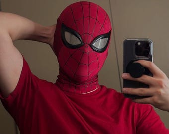 May include: A red Spider-Man mask featuring a black web pattern and large, white, almond-shaped eye lenses with thick black frames. The mask is made of a textured fabric and is paired with a plain red short-sleeved t-shirt.