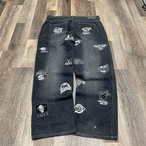 May include: Black denim jeans featuring a variety of white embroidered NBA team logos and basketball graphics scattered across the front. The pants have a relaxed fit, a button fly, and a dark wash finish with visible stitching throughout the fabric.