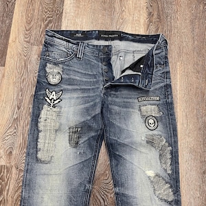 May include: Blue denim jeans with a distressed finish, featuring multiple patches including a skull, wings, and the text "AFFLICTION". The jeans have a button fly, a black waistband label reading "BLACK PREMIUM", and several frayed, stitched-over patches.