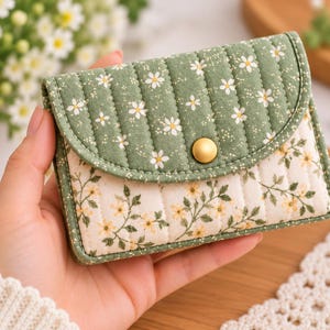 May include: A quilted fabric wallet featuring a sage green flap with a white daisy print and a cream-colored body with a delicate yellow floral vine pattern. The wallet is secured with a single matte gold-toned metal snap button closure.