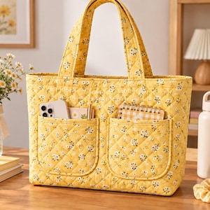 May include: A yellow quilted tote bag featuring a small white floral pattern. The bag has two top handles and two large exterior front pockets. The pockets contain a smartphone, a small notebook, a pen, and a checkered notepad.