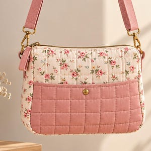 May include: A quilted crossbody bag featuring a cream-colored fabric with a pink and green floral pattern on the upper section and a solid dusty pink quilted pocket on the lower front, secured with a brass-toned snap and an adjustable pink strap.