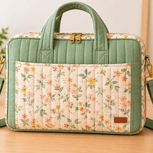 May include: A quilted laptop bag featuring a sage green top and side panels with a cream-colored front pocket adorned with a small orange and yellow floral print. The bag includes two top handles, a detachable shoulder strap, and gold zippers.