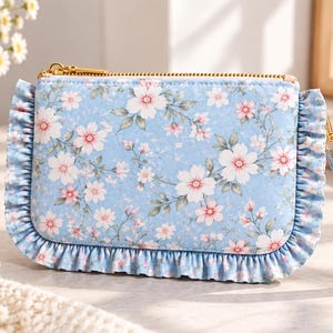 May include: A light blue rectangular pouch featuring a floral pattern of white and pink blossoms with green leaves. The pouch has a gold-tone zipper closure, a matching wrist strap, and ruffled fabric trim along the left and right sides.