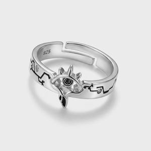May include: A silver-colored open ring featuring a central eye-shaped motif with black enamel detailing and small embedded stones. The band is engraved with the number 925 and a jagged line pattern that extends around the circumference of the ring.
