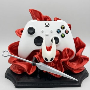 Peut inclure: Une manette sans fil Xbox blanche repose contre une base rouge aux volants rappelant du tissu. Un masque de personnage blanc à cornes avec des fentes oculaires noires est placé devant la manette, tenant une longue arme argentée en forme d'aiguille.
