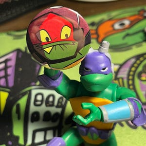 May include: A plastic action figure of a green turtle character with a purple mask, holding a circular button pin featuring a graphic illustration of a red-masked turtle face with yellow eyes and a green mouth area, set against a dark background.
