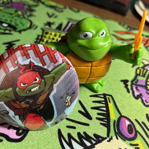May include: A round pinback button featuring a graphic illustration of a green turtle character with a red mask and the text HHHHH, positioned next to a small green and orange plastic turtle figurine holding a yellow weapon accessory.
