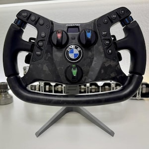 Könnte beinhalten: Ein schwarzes Fanatec-Rennlenkrad aus Kohlefaser mit einem zentralen BMW-Logo, mehreren runden Tasten mit verschiedenen Funktionsbeschriftungen und drei farbigen Drehreglern in Rot, Blau und Grün, montiert auf einem grauen Ständer.