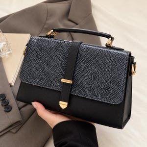 May include: A structured handbag featuring a black base and a grey snakeskin-patterned flap. The bag includes a top handle, a vertical black strap with a gold-toned metal accent, and gold-toned hardware attachments for a shoulder strap.