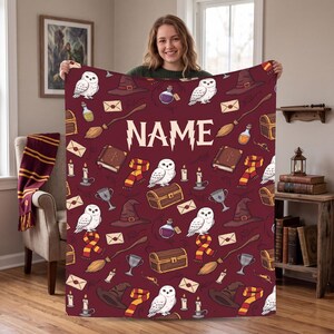 May include: A maroon throw blanket featuring a repeating pattern of white owls, brown wizard hats, striped scarves, wooden wands, potion bottles, envelopes, and treasure chests. The word NAME is printed in large white letters in the center.