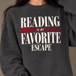 May include: A charcoal gray crewneck sweatshirt featuring the white text READING IS MY FAVORITE ESCAPE with two horizontal red lines between the words READING and IS MY, and between IS MY and FAVORITE.