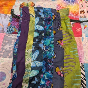 May include: A drawstring pouch constructed from vertical fabric strips featuring various patterns including blue butterflies, geometric shapes, and floral motifs in shades of purple, lime green, and dark blue with a dark green cord closure.