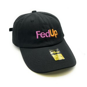 May include: A black baseball cap featuring the embroidered text FedUp in pink and orange letters on the front. A rectangular black and yellow sticker is attached to the brim, displaying the text HATZONE and Official Licensed Product.