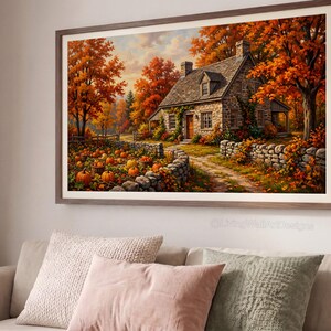Op de afbeelding: Een ingelijste landschaps-muurkunstprint met een stenen huisje omringd door herfstbomen met oranje en rood gebladerte. Een zandpad leidt naar het huis, langs een stenen muur en een pompoenveld vol oranje pompoenen onder een bewolkte lucht.