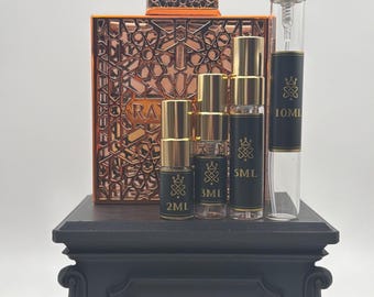 May include: A collection of four clear glass perfume spray vials labeled 2ml, 3ml, 5ml, and 10ml with gold caps, arranged in front of a large square copper-colored bottle with an intricate geometric cutout design and the text Royale Decants.