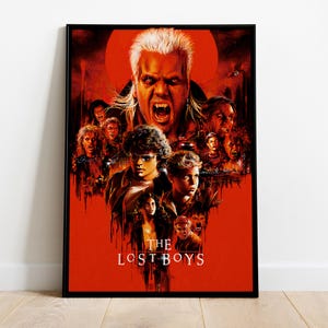 May include: A framed movie poster for The Lost Boys featuring a large central figure with fangs and spiky hair, surrounded by a collage of various characters in a dark, red-toned color palette with the title text The Lost Boys at the bottom.