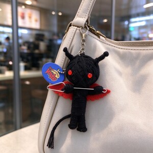 May include: A black yarn-wrapped doll keychain with red bead eyes, red felt wings, and a silver safety pin across its chest. A blue circular tag with the text Voodoo is attached to the metal chain, hanging from a cream-colored handbag.