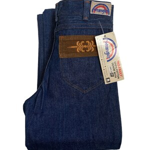 May include: A pair of dark blue denim jeans with a brown corduroy patch on the back pocket. The jeans have a Wrangler label on the back pocket and a tag that reads 'NO-FAULT 100% COTTON DENIM WITH SANFOR-SET'.