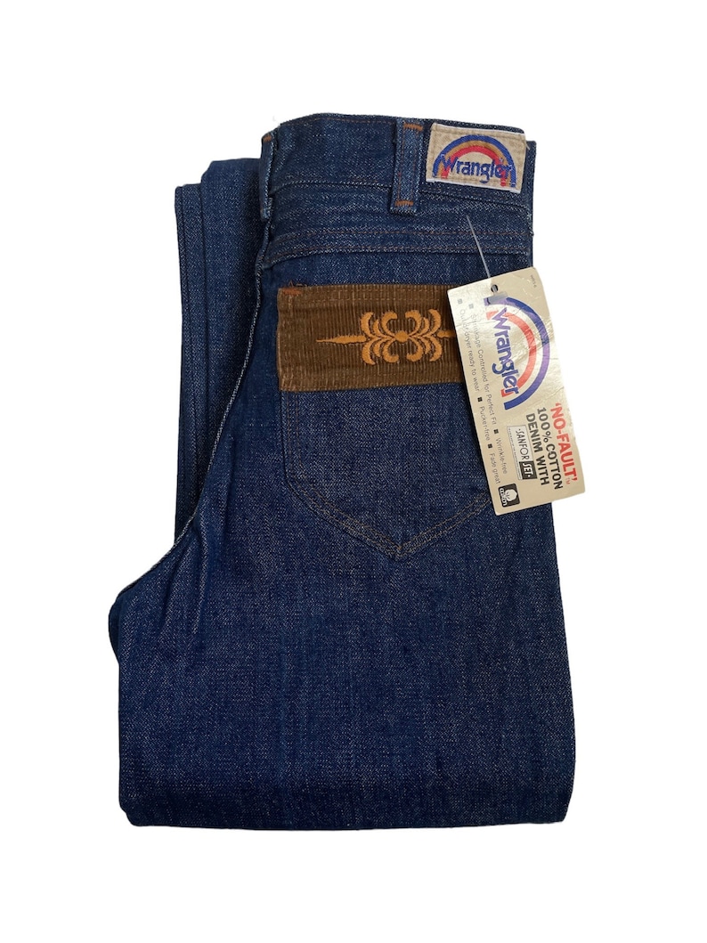 May include: A pair of dark blue denim jeans with a brown corduroy patch on the back pocket. The jeans have a Wrangler label on the back pocket and a tag that reads 'NO-FAULT 100% COTTON DENIM WITH SANFOR-SET'.