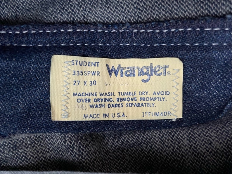 以下が含まれることがあります： 「STUDENT 335SWR 27 X 30」と洗濯表示が書かれた、ラングラーのジーンズのタグのクローズアップ。タグはベージュで白いステッチが施され、ラングラーのロゴは青色です。