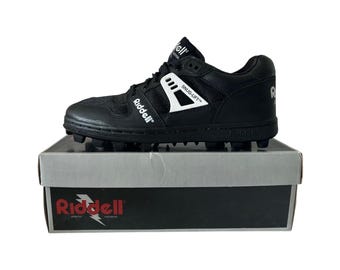 May include: Black Riddell football cleat with white accents, sitting on a gray and black box. The cleat features the Riddell logo and the words "SNUG-LIFT". The box has the Riddell logo and the words "ATHLETIC EQUIPMENT".