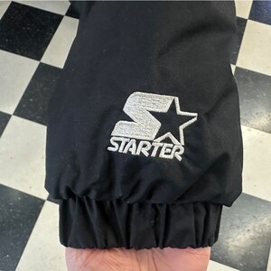 May include: Black jacket sleeve with a white embroidered Starter logo featuring a star. The sleeve has a ribbed cuff. The background is a black and white checkered pattern.