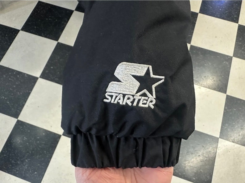 May include: Black jacket sleeve with a white embroidered Starter logo featuring a star. The sleeve has a ribbed cuff. The background is a black and white checkered pattern.