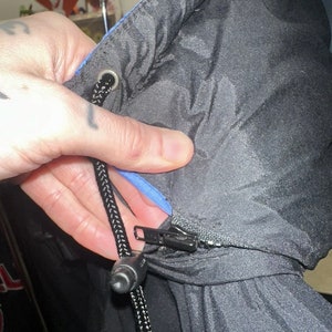 May include: Black puffer jacket with a blue interior lining. The jacket has a black zip closure and a drawstring with a black cord and a plastic stopper. The hood of the jacket is visible.
