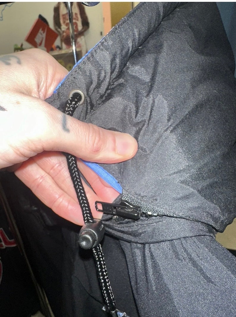 May include: Black puffer jacket with a blue interior lining. The jacket has a black zip closure and a drawstring with a black cord and a plastic stopper. The hood of the jacket is visible.