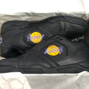 Puede incluir: Zapatillas de baloncesto de cuero negro con el logotipo de Los Angeles Lakers en morado y amarillo. Las zapatillas tienen una suela negra con un patr&oacute;n texturizado.