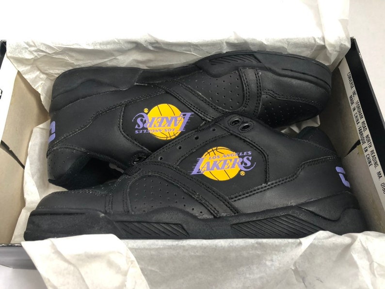 Puede incluir: Zapatillas de baloncesto de cuero negro con el logotipo de Los Angeles Lakers en morado y amarillo. Las zapatillas tienen una suela negra con un patr&oacute;n texturizado.