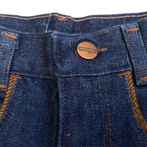 May include: Close-up of the waistband of a pair of blue denim jeans with a brown leather patch that says "Wrangler" on it.