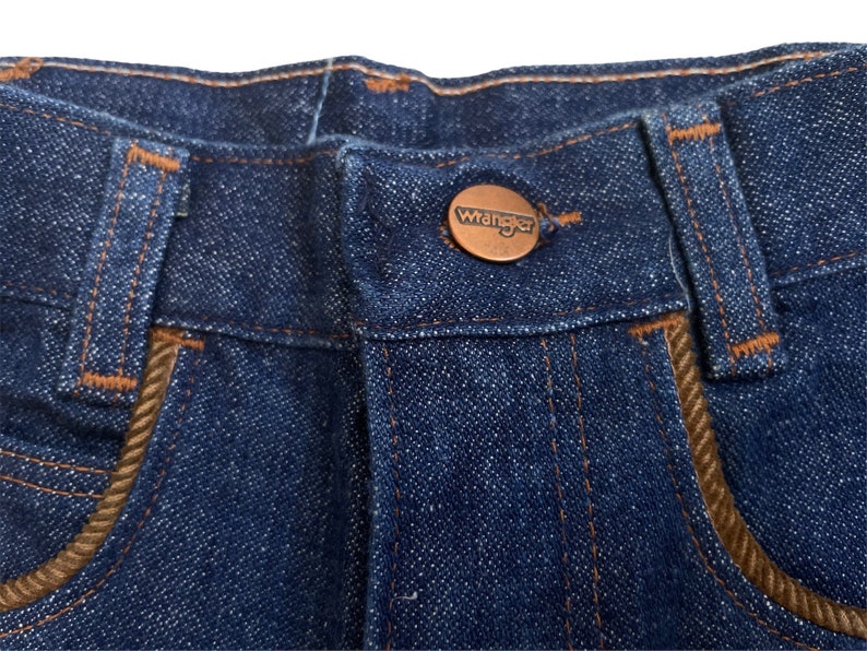 May include: Close-up of the waistband of a pair of blue denim jeans with a brown leather patch that says "Wrangler" on it.