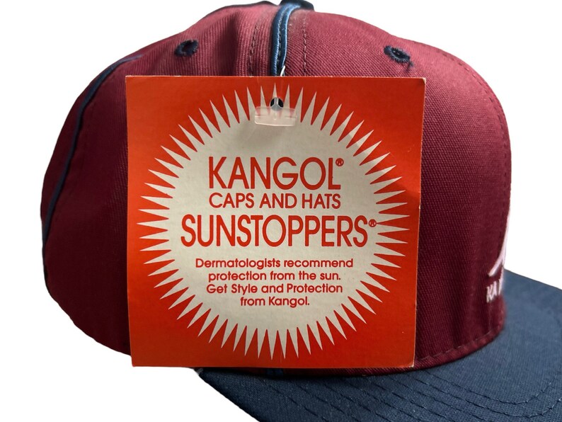 May include: A red and navy blue baseball cap with a white Kangol logo and the text "Caps and Hats Sunstoppers". The tag also includes the text "Dermatologists recommend protection from the sun. Get Style and Protection from Kangol."